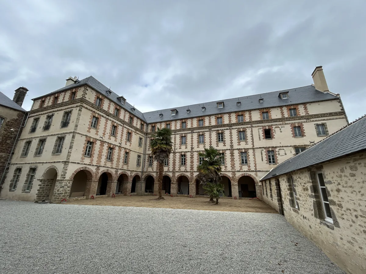 Façades enduites, encadrements en pierre et brique et arcades autour d’une cour.
